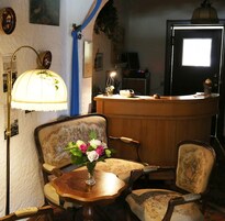 Um quarto com uma receção de madeira, uma cadeira vintage com estofos florais, uma pequena mesa redonda com um vaso de flores e uma luminária de parede.