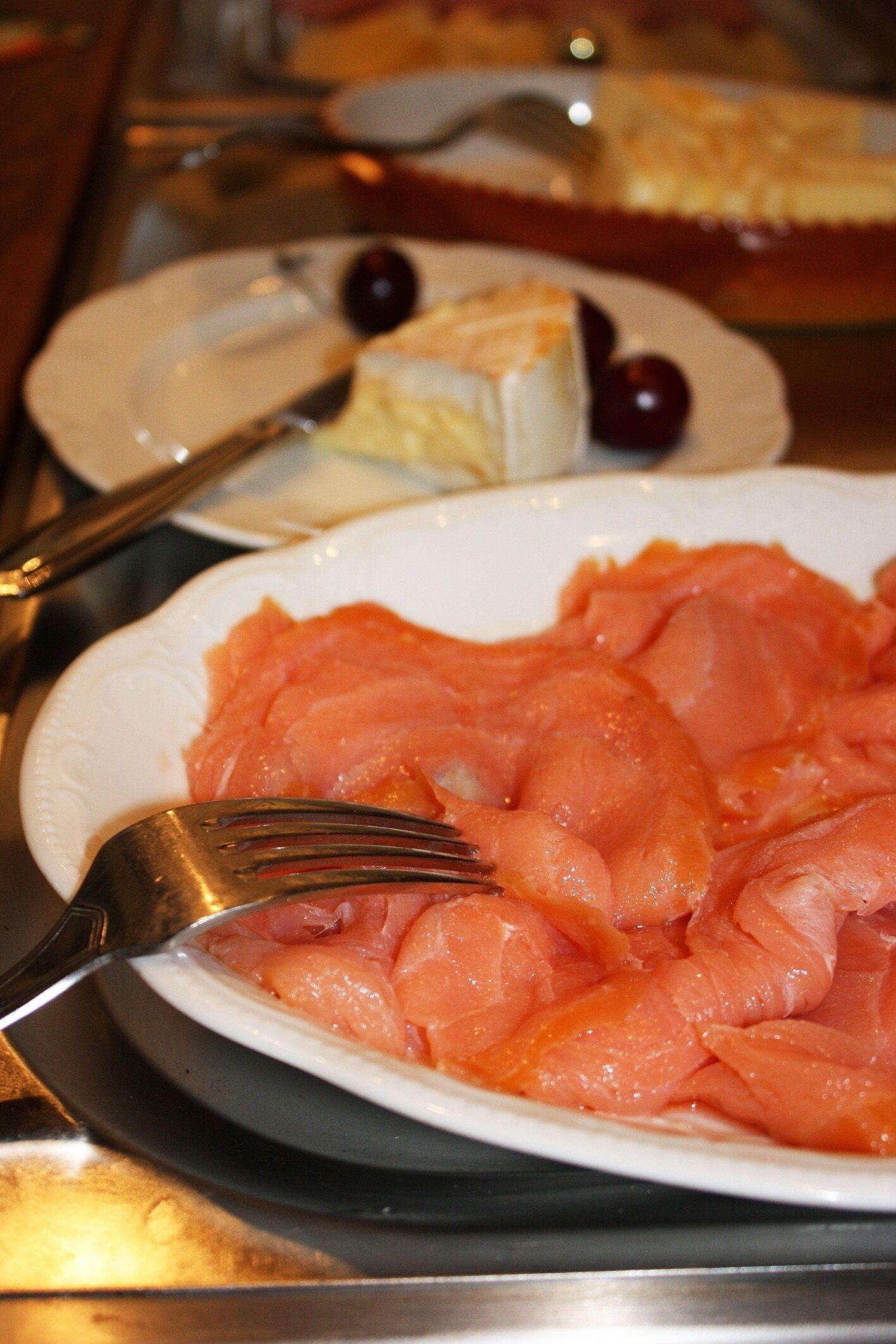 A plate of smoked salmon with a fork on it, a plate with a slice of cheese and a cherry, and a pie in the background.