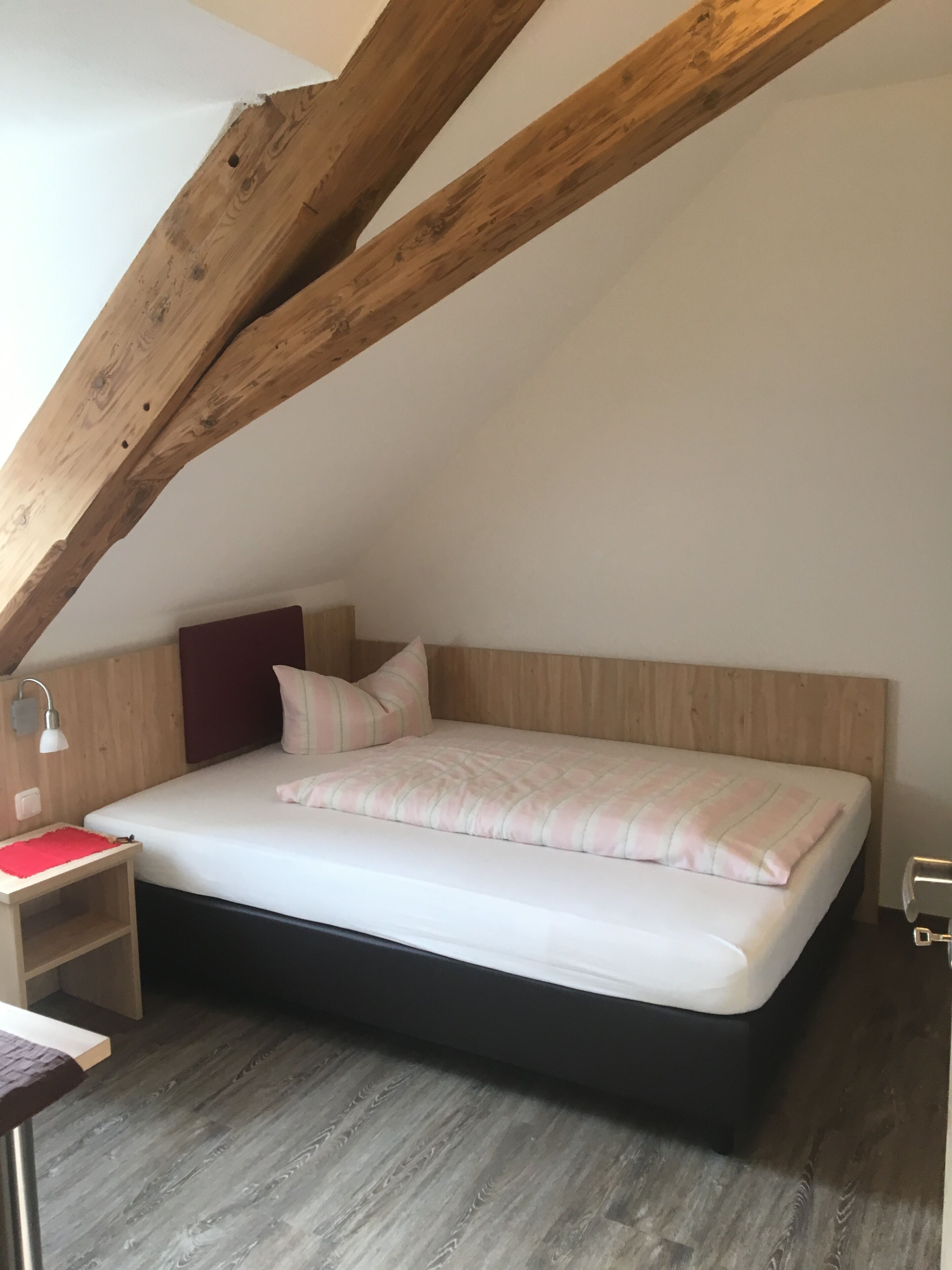 A bedroom with a sloped ceiling, a bed with a white mattress and striped bedding, a bedside table with a lamp, and a wooden headboard.