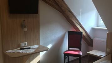 A small, cozy room with a sloped ceiling, a wooden desk with a chair, a bed, and a television mounted on the wall.