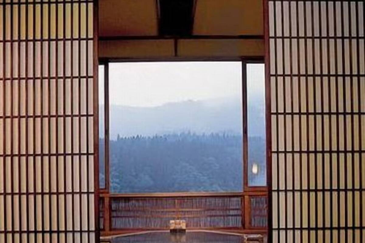 A room with a wooden table and chairs, a view of a forest through a large window, and a traditional Japanese sliding door.