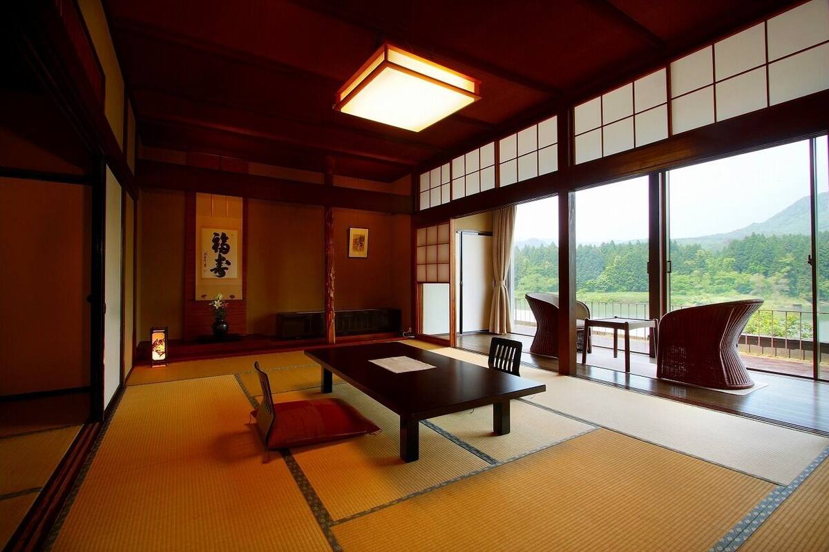A traditional Japanese room with tatami flooring, a low table, a chair, and a view of a forested mountain.