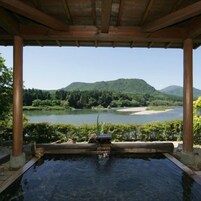 Zona de aguas termales con una pérgola de madera, vista a un lago y una montaña al fondo.