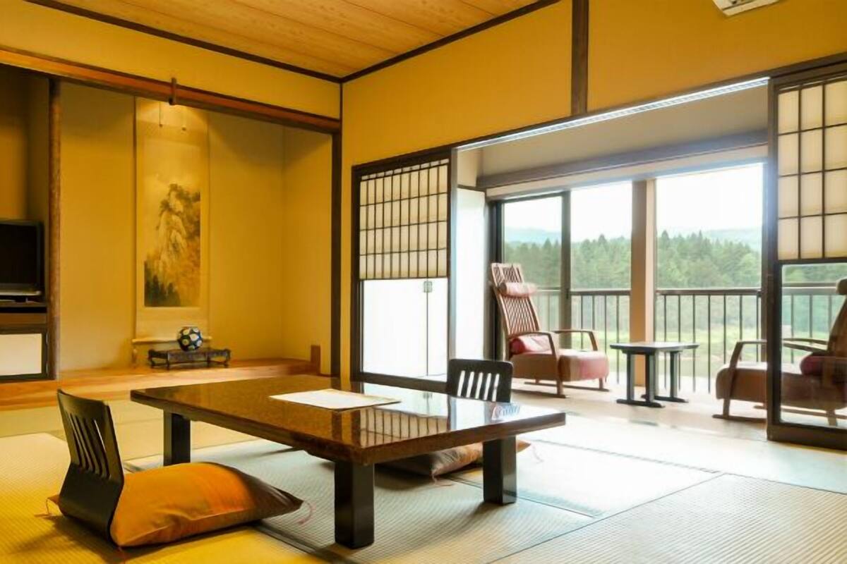 A traditional Japanese-style room with tatami flooring, a low table, chairs, and a sliding door leading to a balcony with a view of the forest.