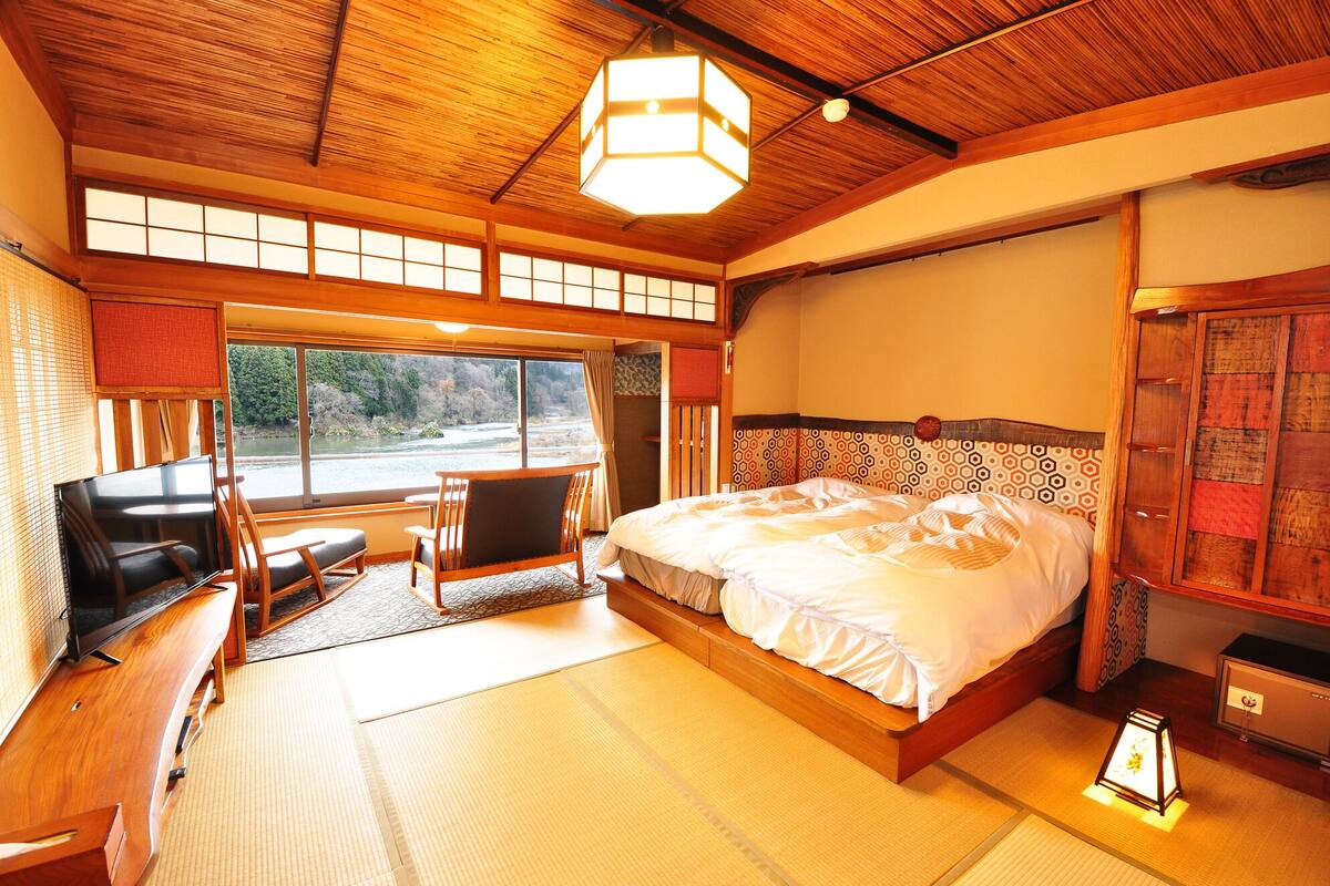 A traditional Japanese-style room with tatami flooring, a bed with a patterned headboard, a television, and a view of a river.