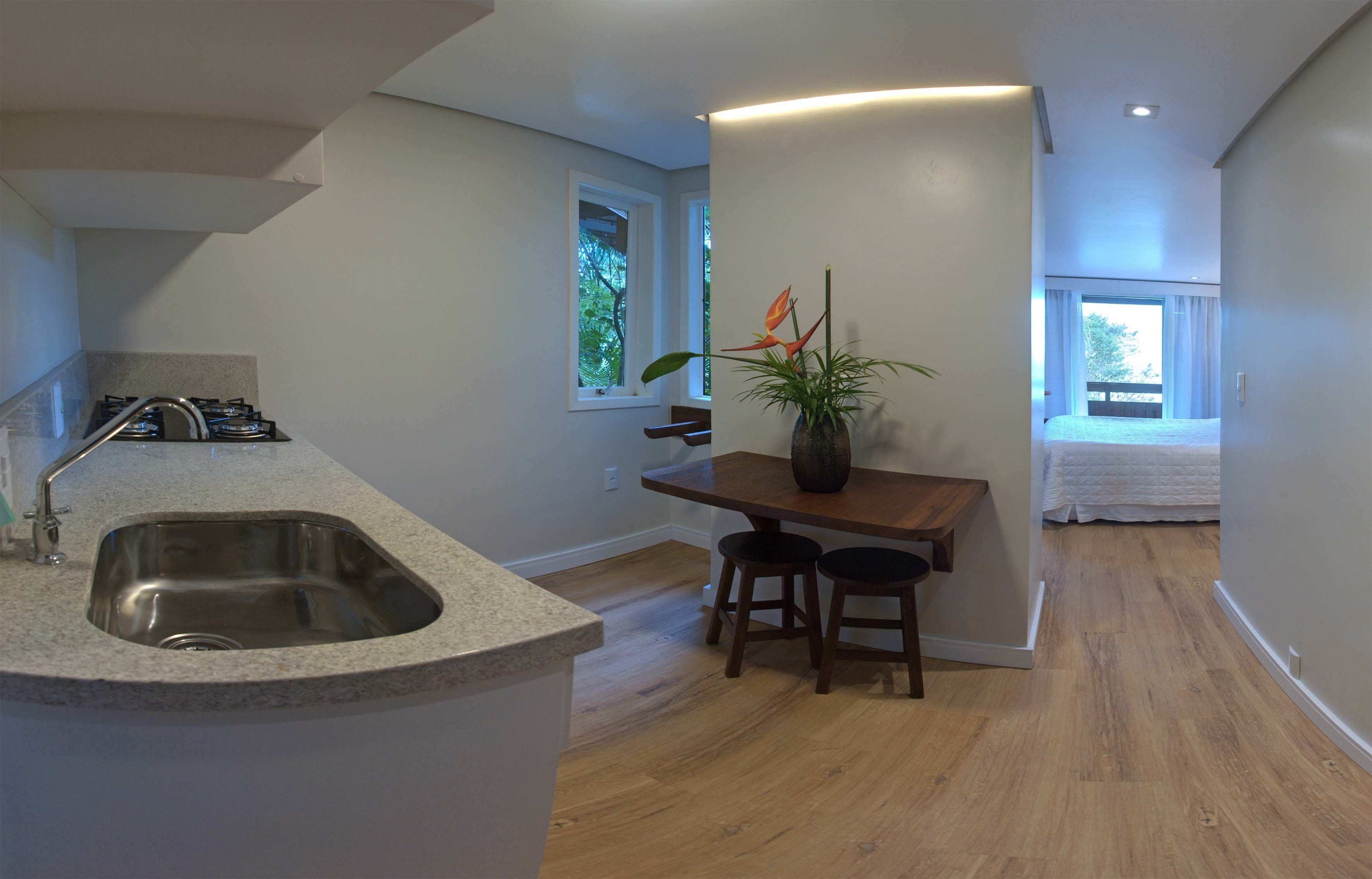 A modern kitchen with a granite countertop, stainless steel sink, and wooden stools. There is a dining table with a vase of flowers and a window with curtains.