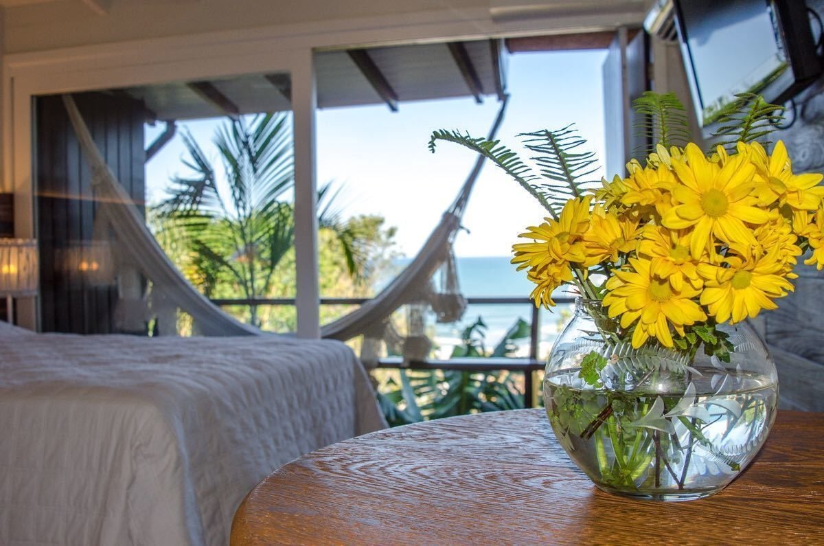 A room with a bed, a round table with a vase of yellow flowers, and a view of the sea through large windows.