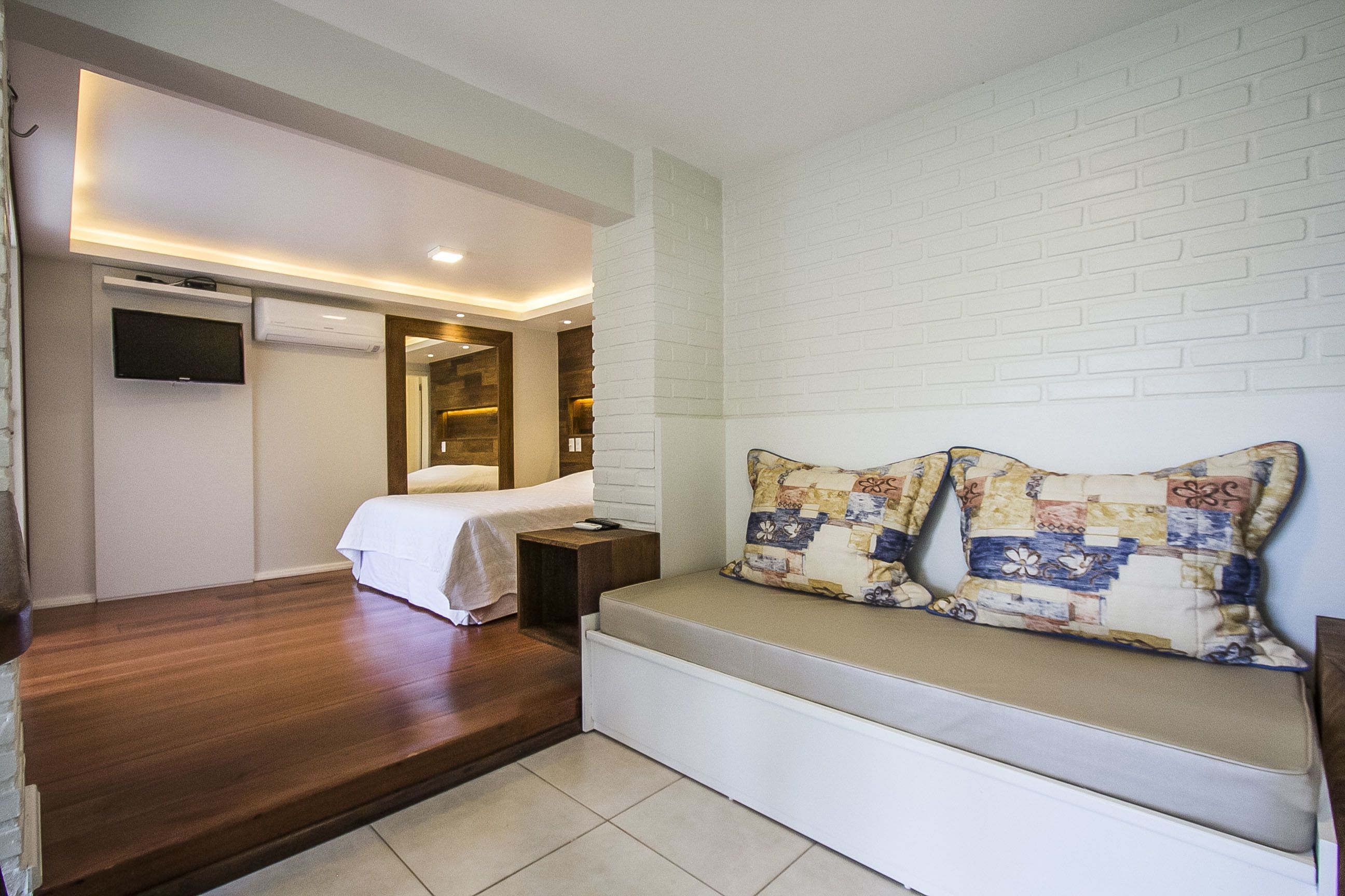 A hotel room with a bed, a bench with a quilted pillow, a flat-screen TV, and a wall-mounted air conditioner.
