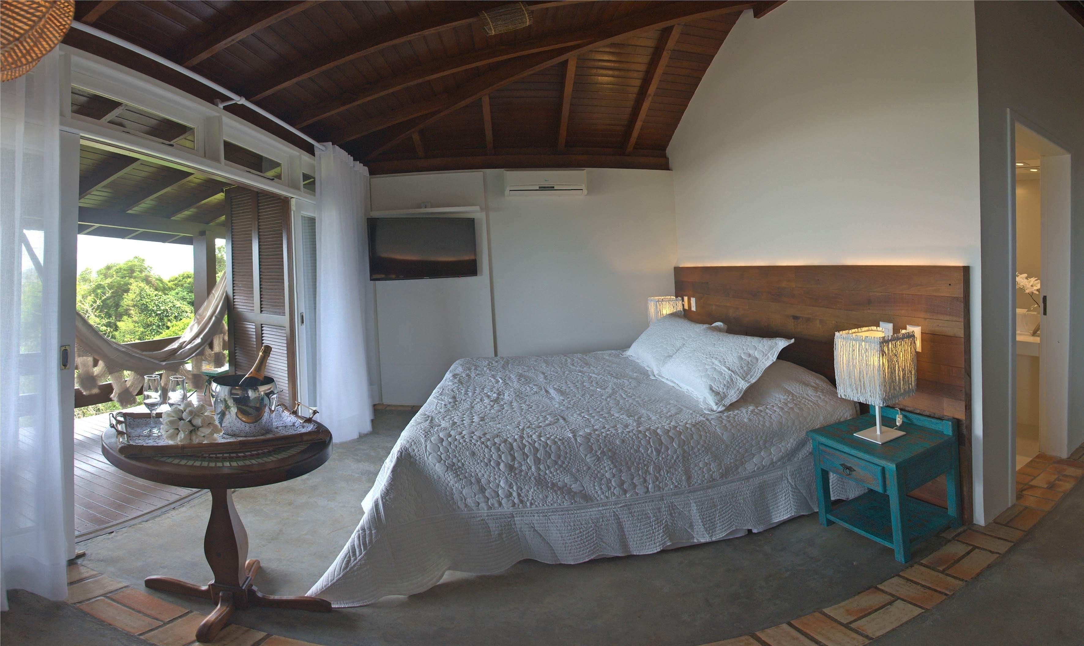 A bedroom with a large bed, a wooden headboard, a nightstand with a lamp, and a view of greenery outside.