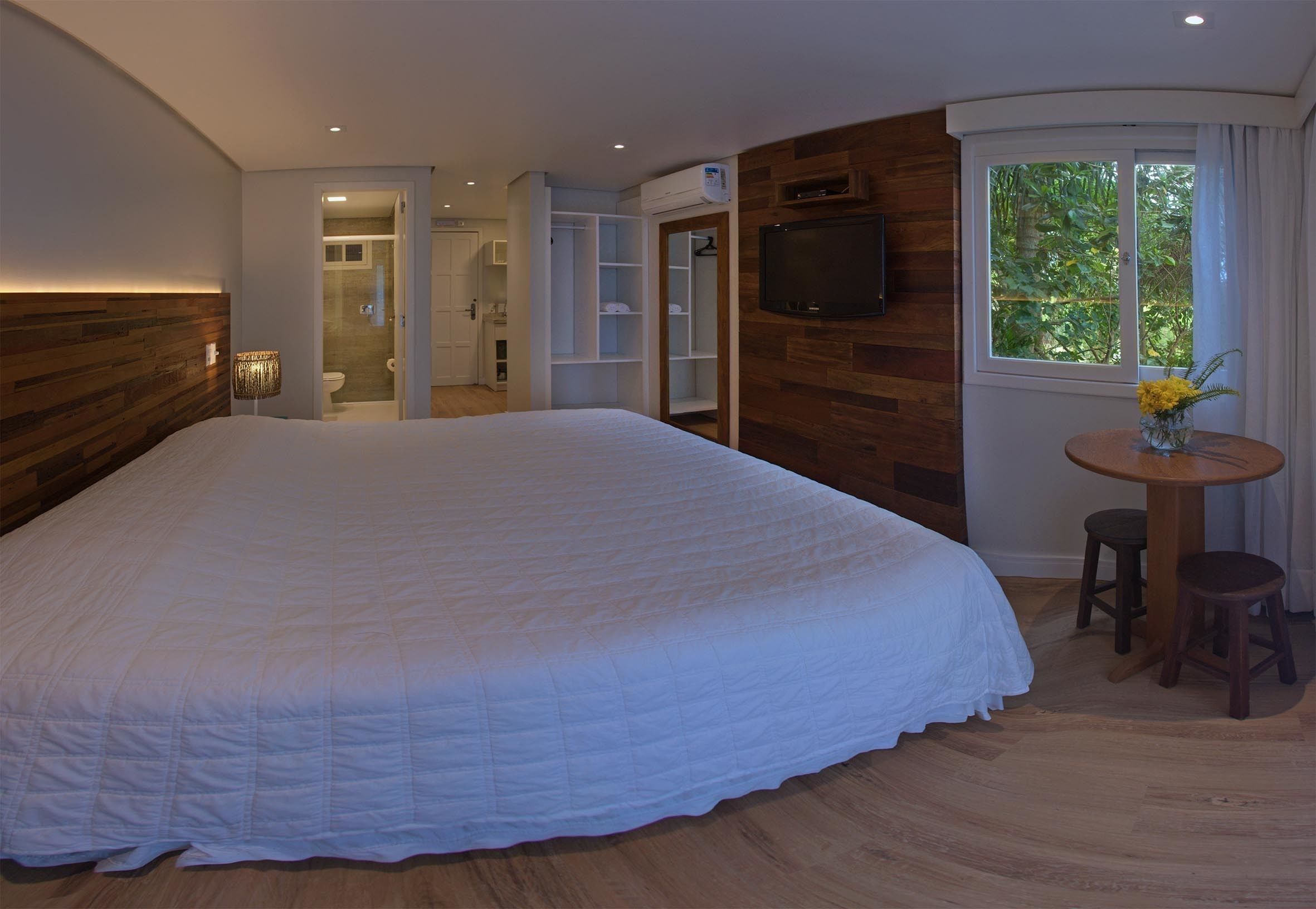 Un dormitorio con una cama grande, televisión montada en la pared, cabecera de madera, ventana con vistas a la vegetación y una pequeña mesa con taburetes.