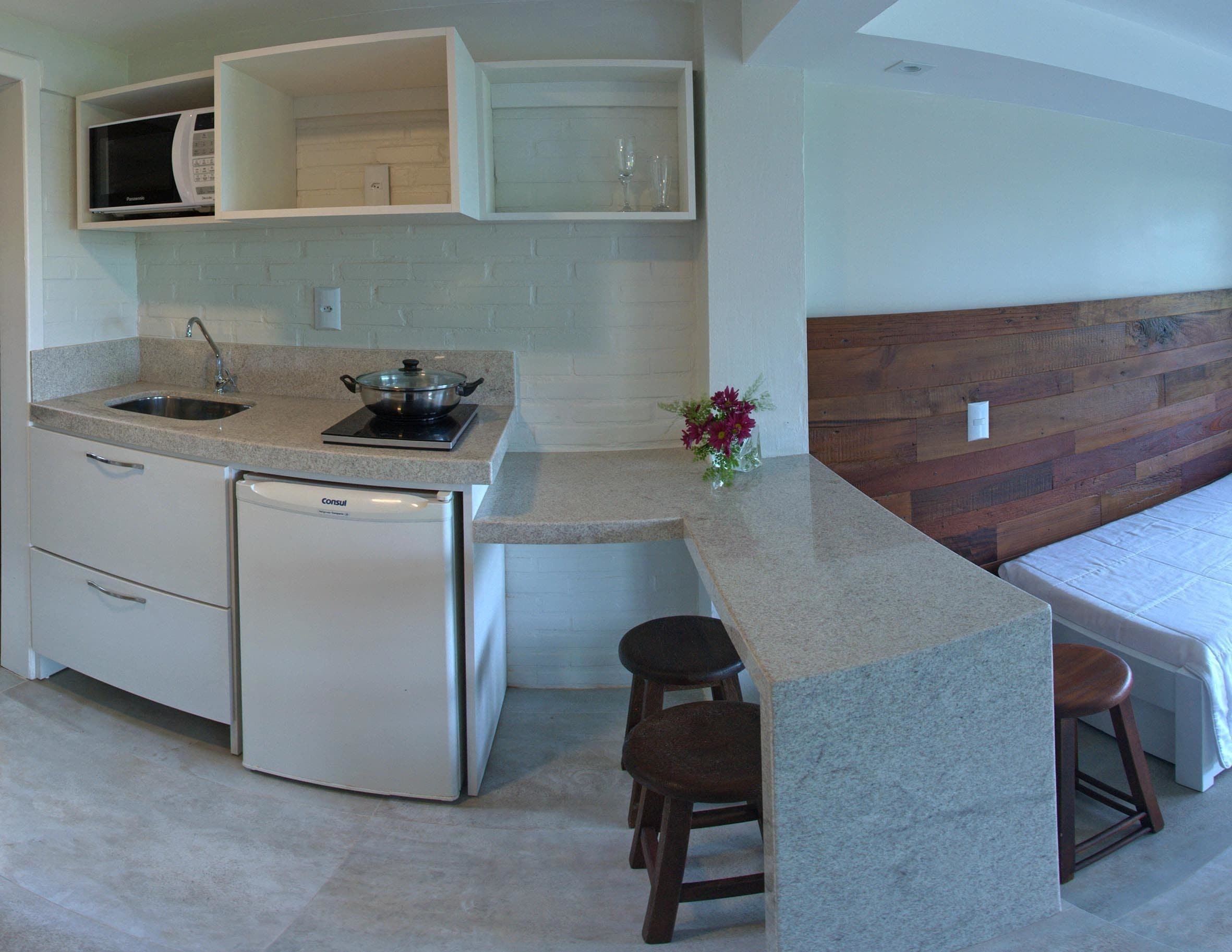 A compact kitchen with a built-in microwave, a sink, and a small refrigerator. There are stools and a wooden headboard in the adjacent sleeping area.