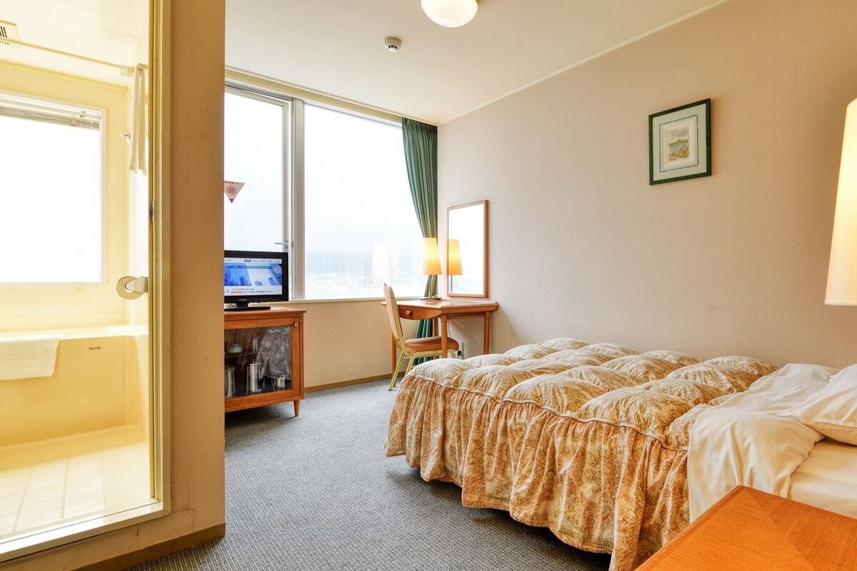 A hotel room with a bed, a desk with a chair, a television, and a window with curtains.