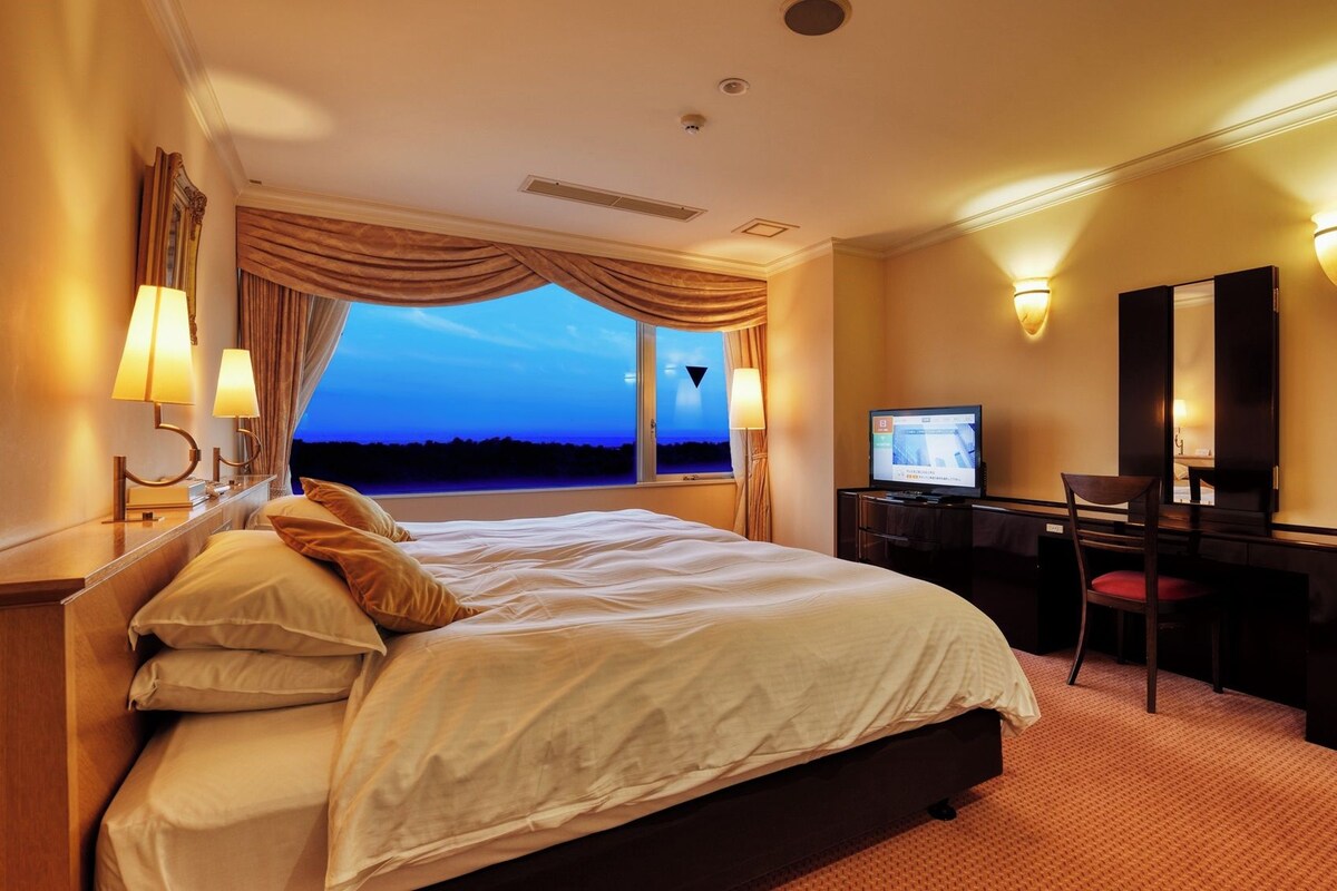 A hotel room with a large bed, a desk with a computer, a TV, and a view of the evening sky.