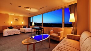 A hotel room with two beds, a sofa, a small table, and a view of the city at dusk. - Plaza Hotel Mutsu (Mutsu)