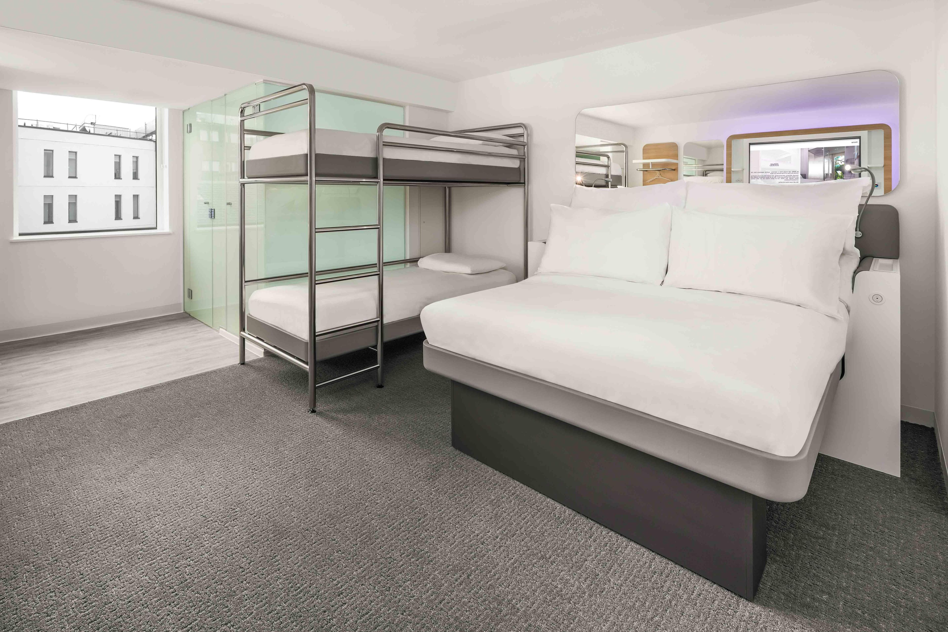 A compact hotel room with bunk beds, a large bed, a mirror, and a window with a view of buildings.