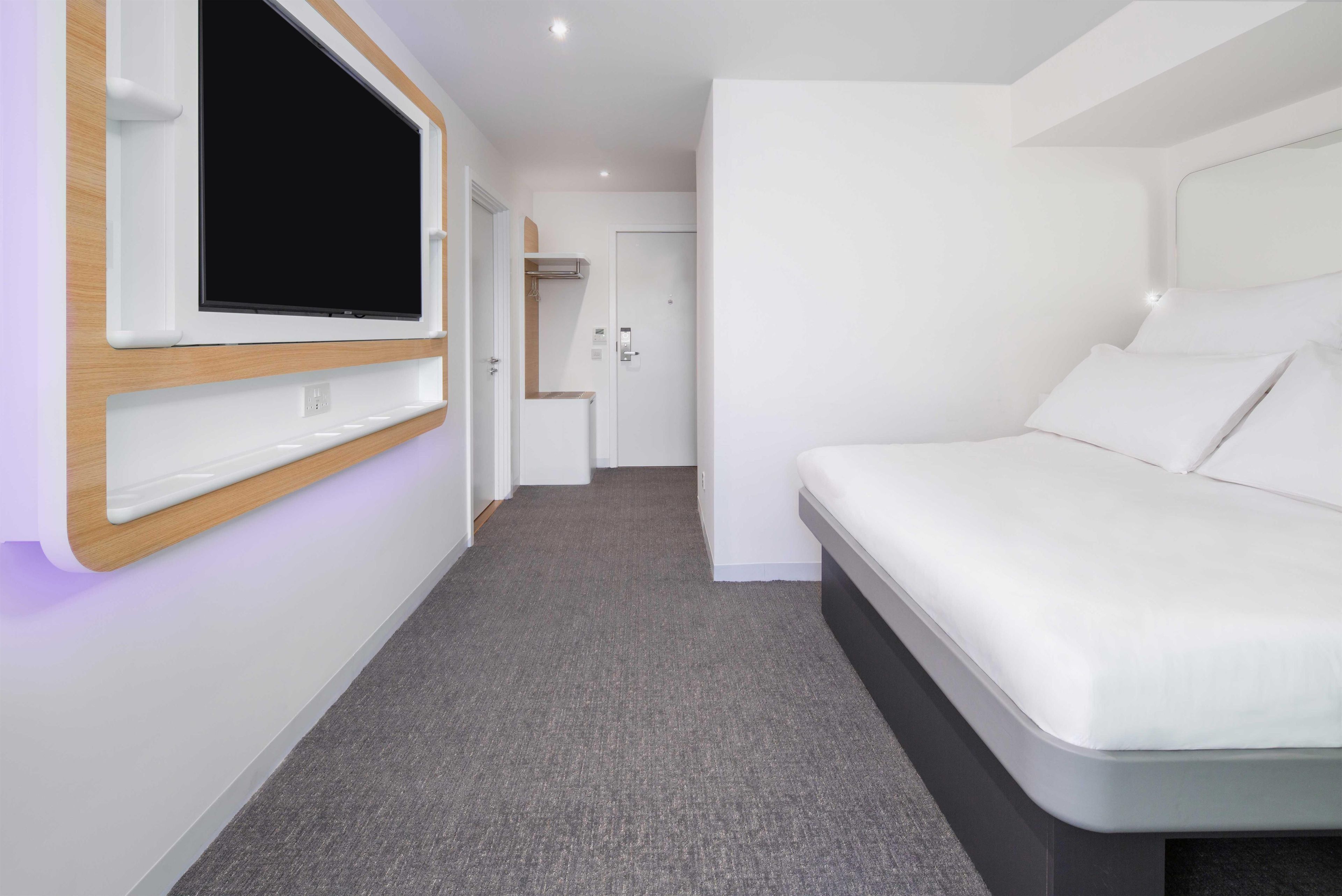 Quarto de hotel moderno com televisão de tela plana na parede, cama com roupa de cama branca e piso coberto por carpete cinza.