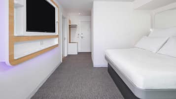 Quarto de hotel moderno com televisão de tela plana na parede, cama com roupa de cama branca e piso coberto por carpete cinza.