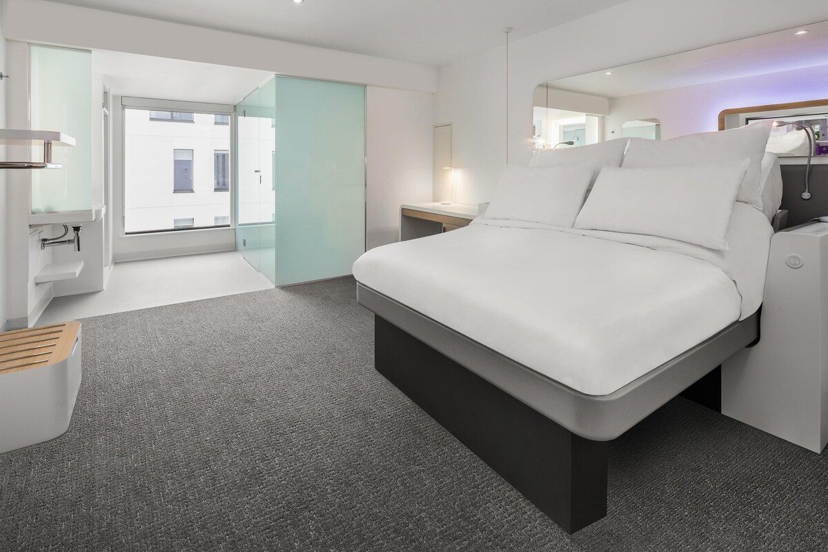 A modern hotel room with a bed, bedside table, and a bathroom visible through an open door.