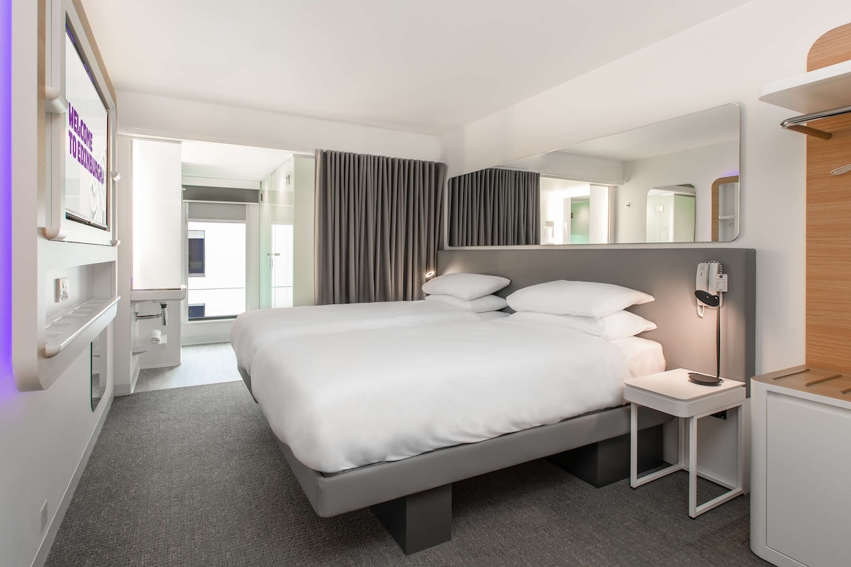 A modern hotel room with a large bed, bedside tables, a mirror, and a view of the outside.