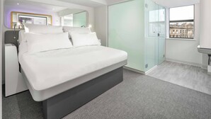 A modern bedroom with a large bed, a glass wardrobe, and a view of buildings through a window.
