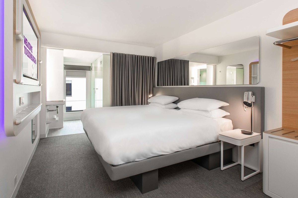 A modern hotel room with a large bed, bedside tables, a mirror, and a view of the outside.