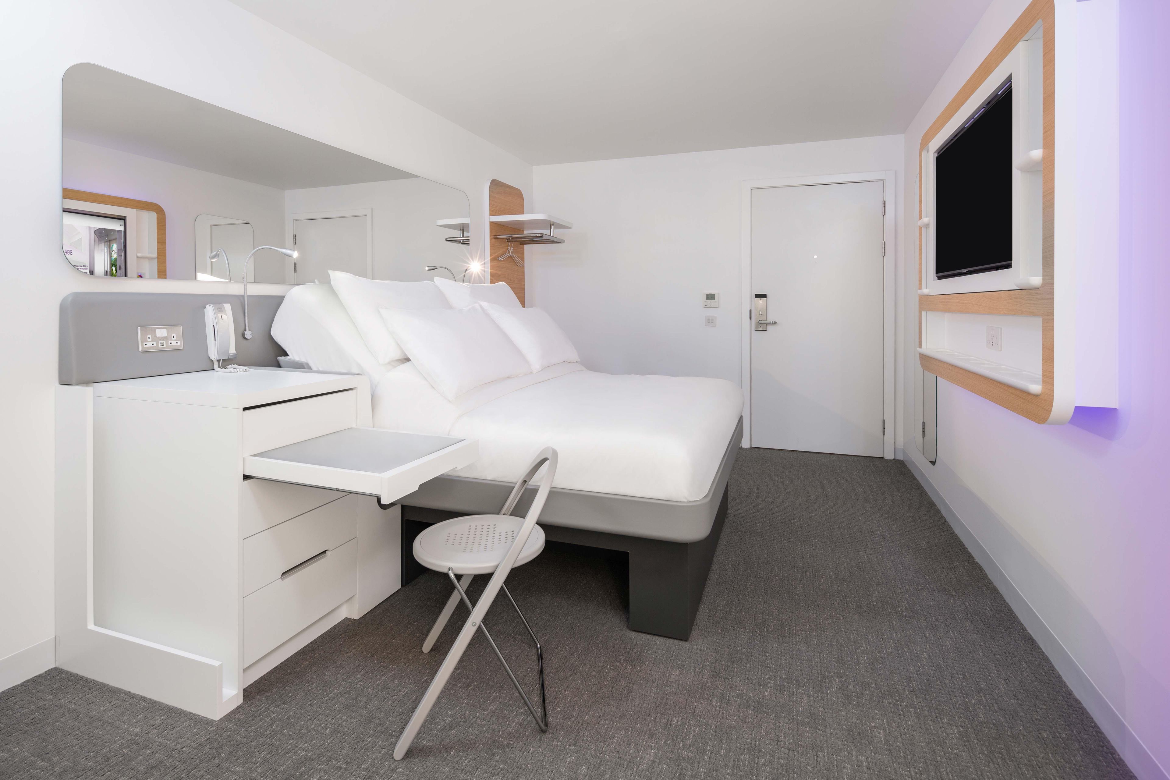 A modern hotel room with a bed, a desk, a chair, a TV, and a mirror.