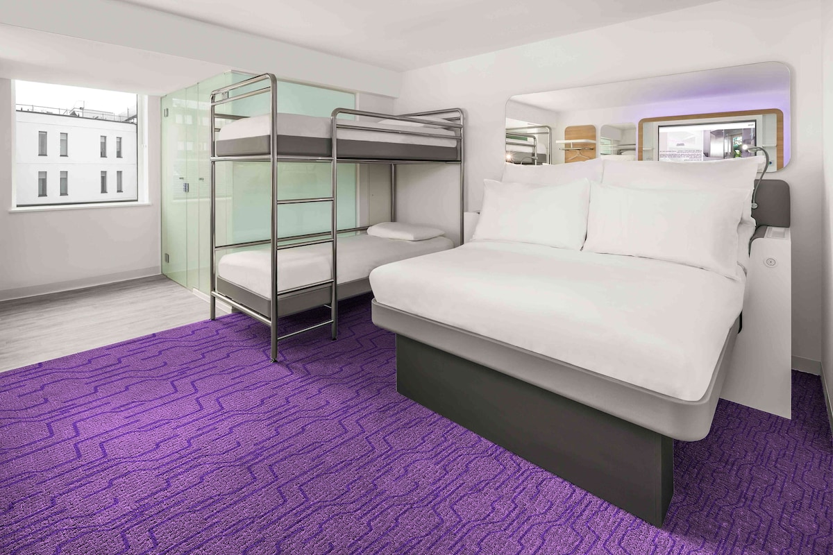 A hotel room with bunk beds, a large bed, a purple carpet, a window, and a mirror.
