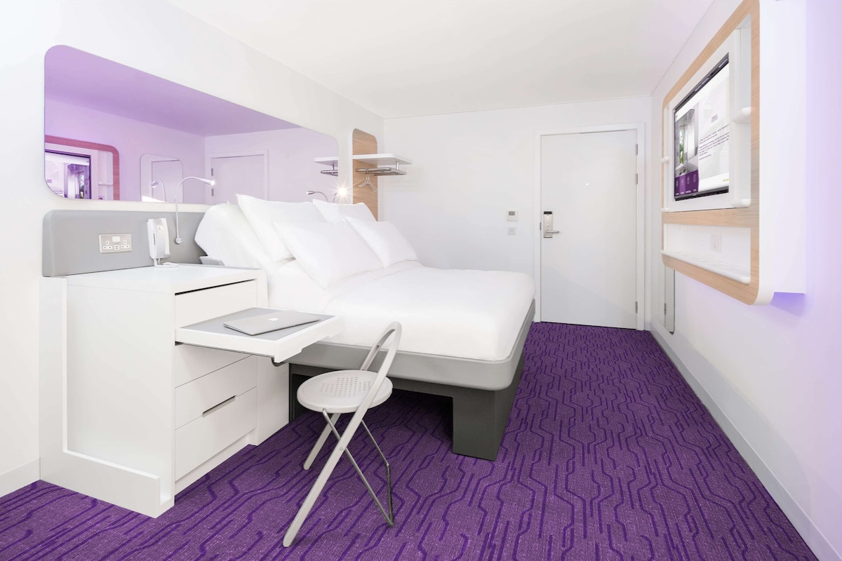 A modern hotel room with a bed, desk, chair, and a small table.