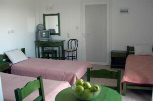 Una habitación sencilla con dos camas, un escritorio con computadora y espejo, y una mesa con un bol de manzanas.