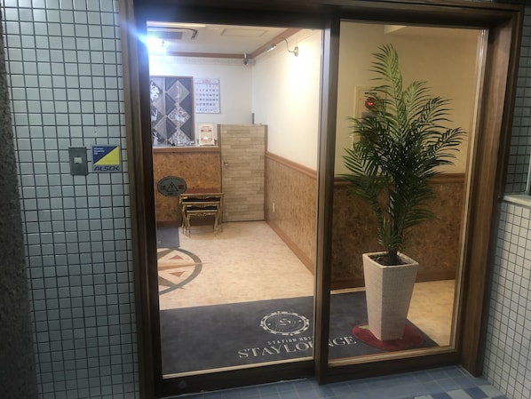 A hotel entrance with a reception desk, a small table, a potted plant, and a doormat with the hotel's name.