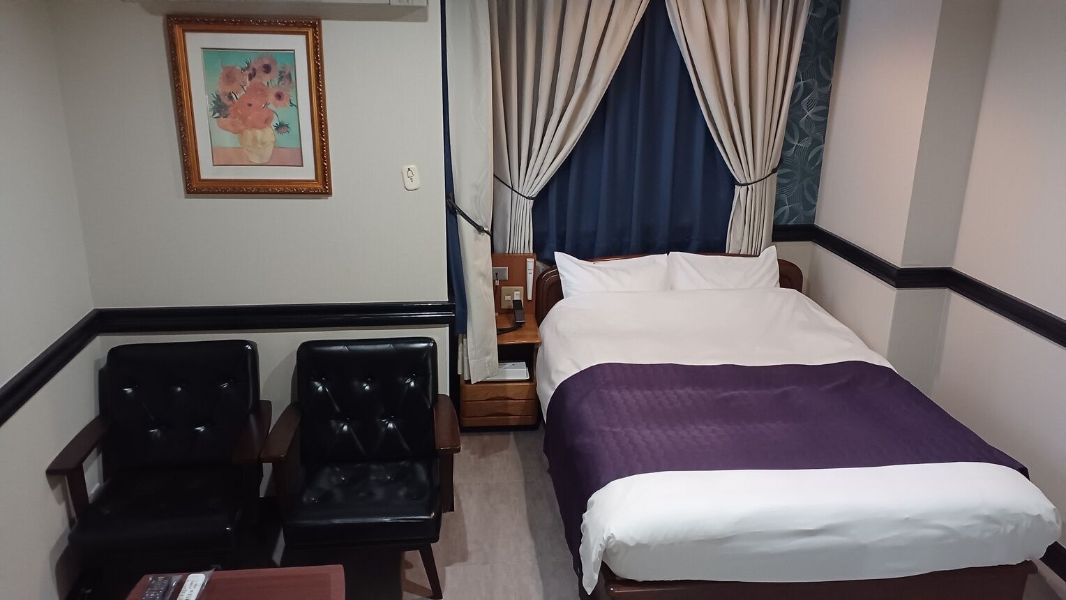 Una habitación de hotel con cama, dos sillas, una mesita de noche, un cuadro y cortinas.