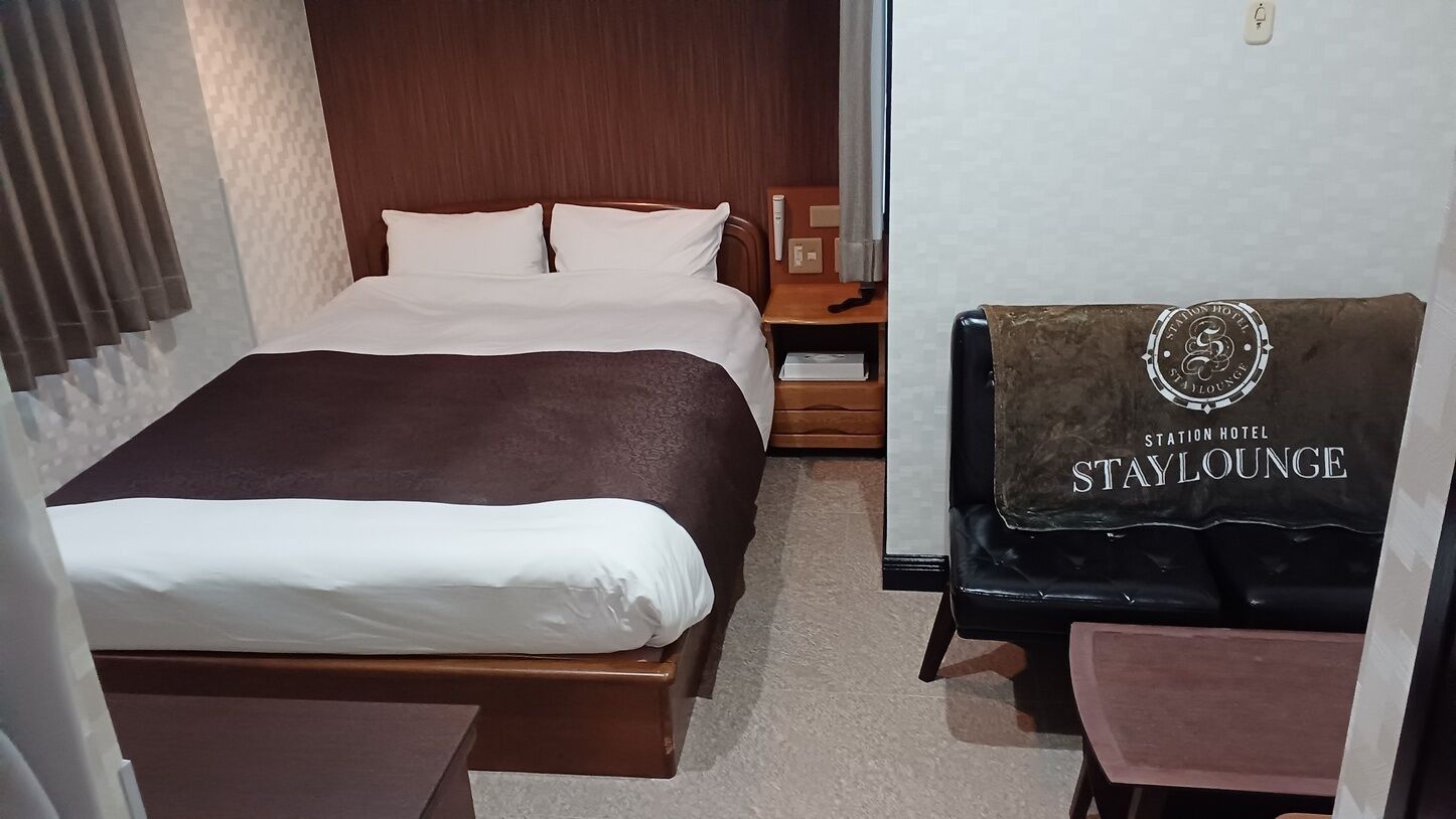 Habitación de hotel con cama, mesita de noche, silla y ventana con cortinas. Se observa un cartel con la inscripción 'Station Hotel Stay Lounge'.
