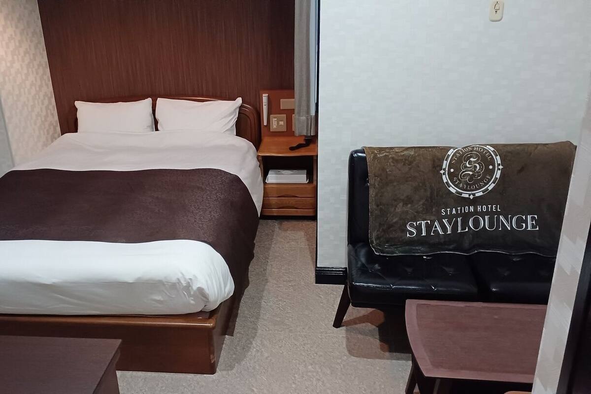 Habitación de hotel con cama, mesita de noche, silla y ventana con cortinas. Se observa un cartel con la inscripción 'Station Hotel Stay Lounge'.