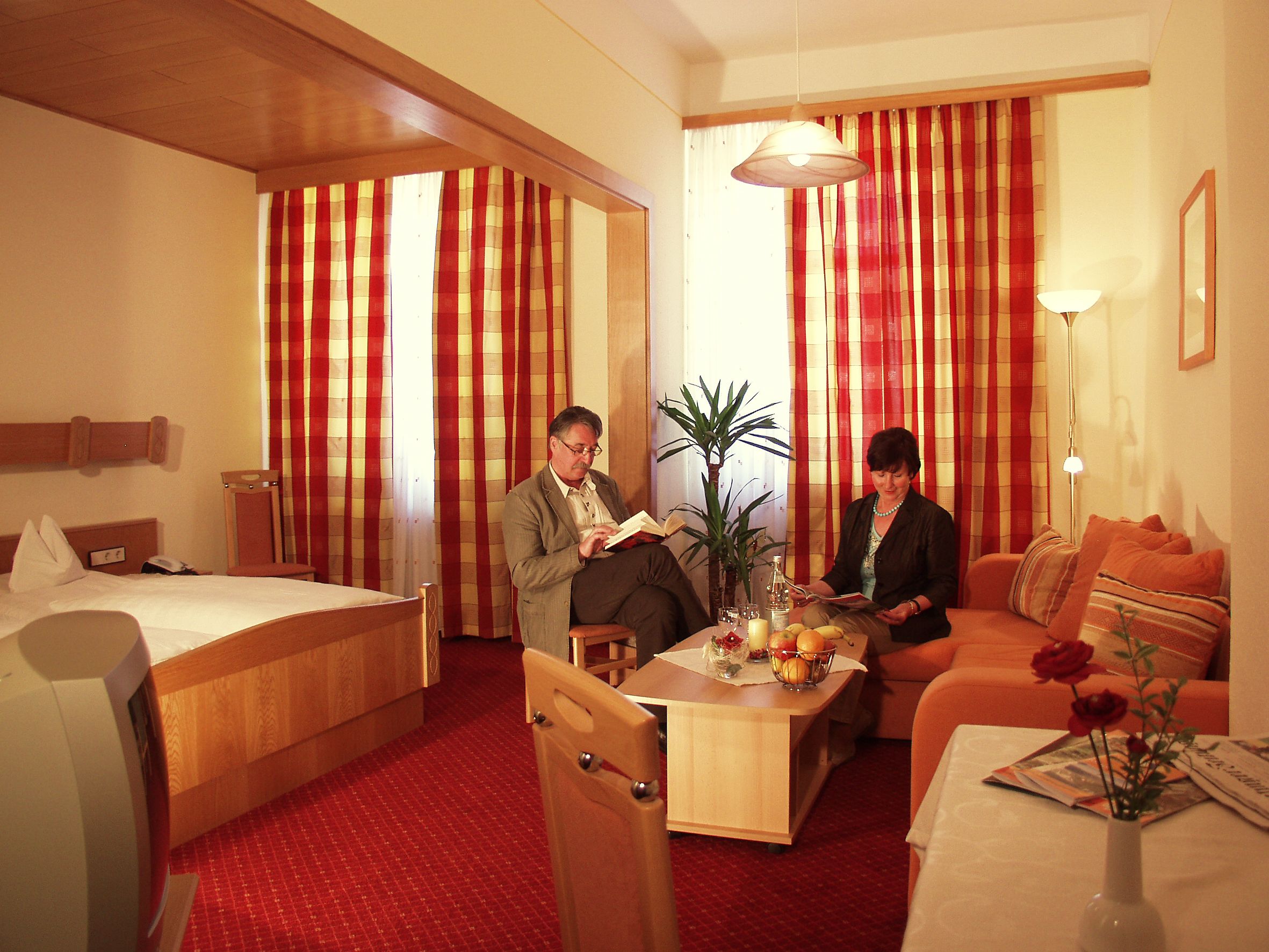 A hotel room with a bed, a sofa, a dining table with a vase of flowers, and a potted plant.