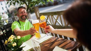 Zwei Personen stoßen mit Biergläsern an einem Holztisch an, auf dem eine Pflanze in einer Vase steht.
