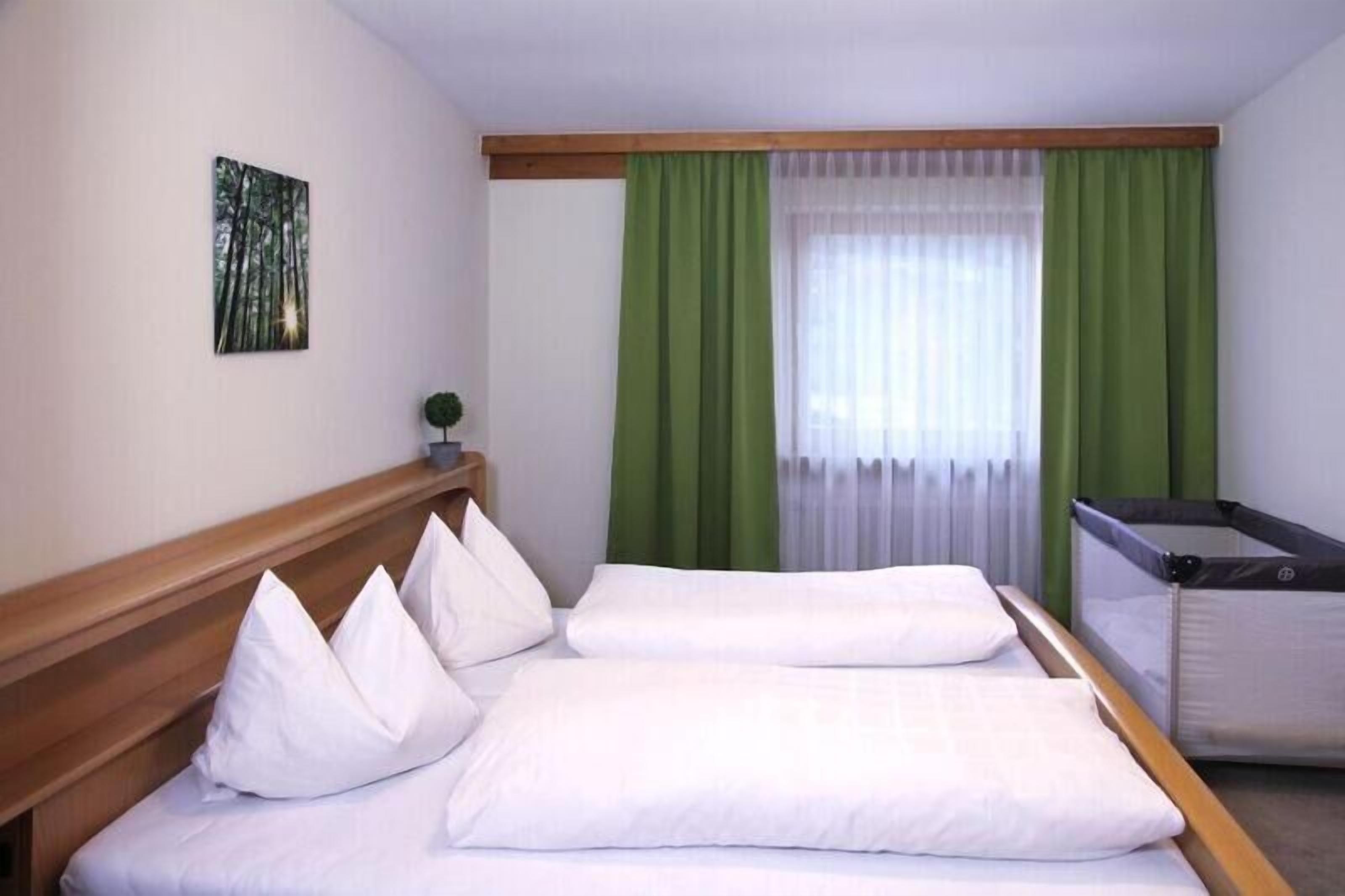 Habitación de hotel con dos camas, cortinas verdes, una ventana con cortinas blancas translúcidas y una pequeña planta en la mesita de noche.