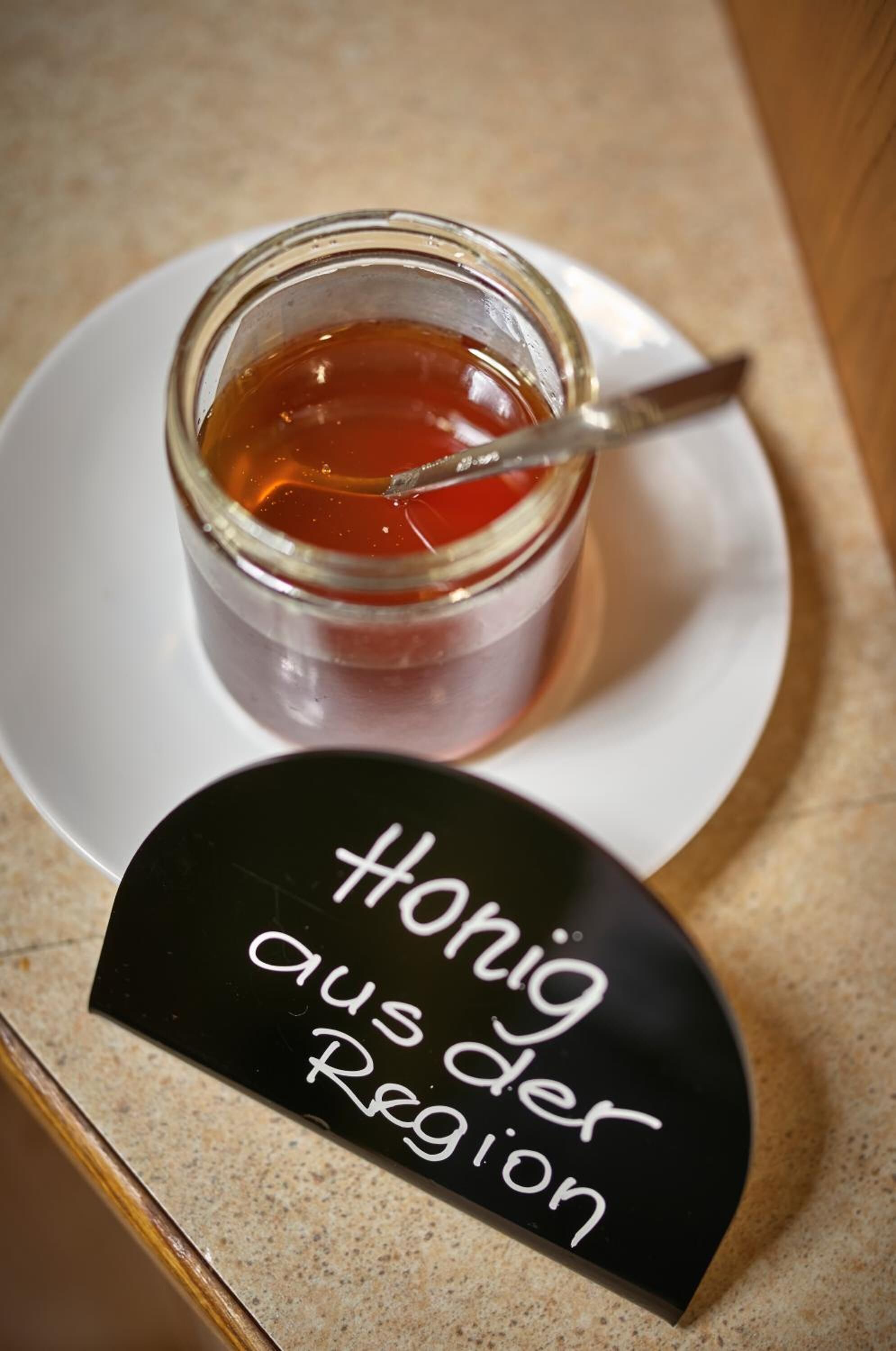 A glass jar of honey with a spoon, placed on a white plate on a countertop.
