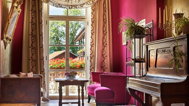 Ein Zimmer mit einem Klavier, einem roten Sessel, einer Bank, einem Tisch mit einer Vase und einem Fenster mit Blick auf einen Garten.
