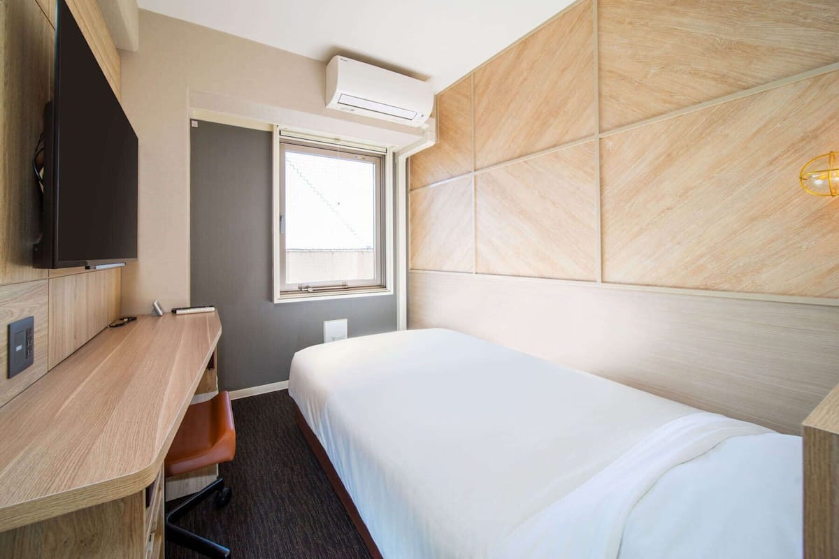 A hotel room with a bed, a desk, a chair, a television, a window, and an air conditioning unit.