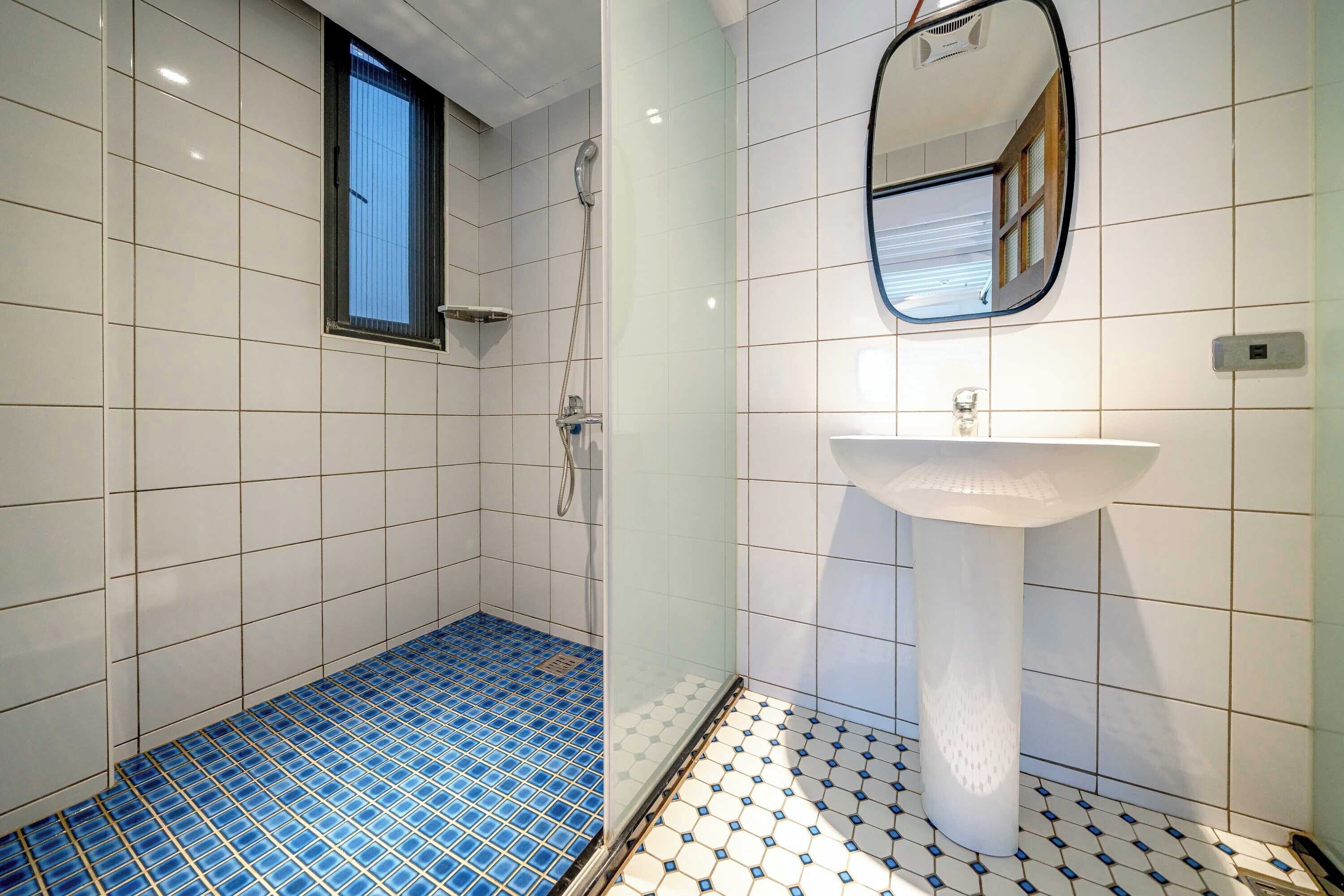 Une salle de bain avec des murs recouverts de carreaux blancs, un sol à motifs bleus et blancs, une douche, un lavabo et un miroir.