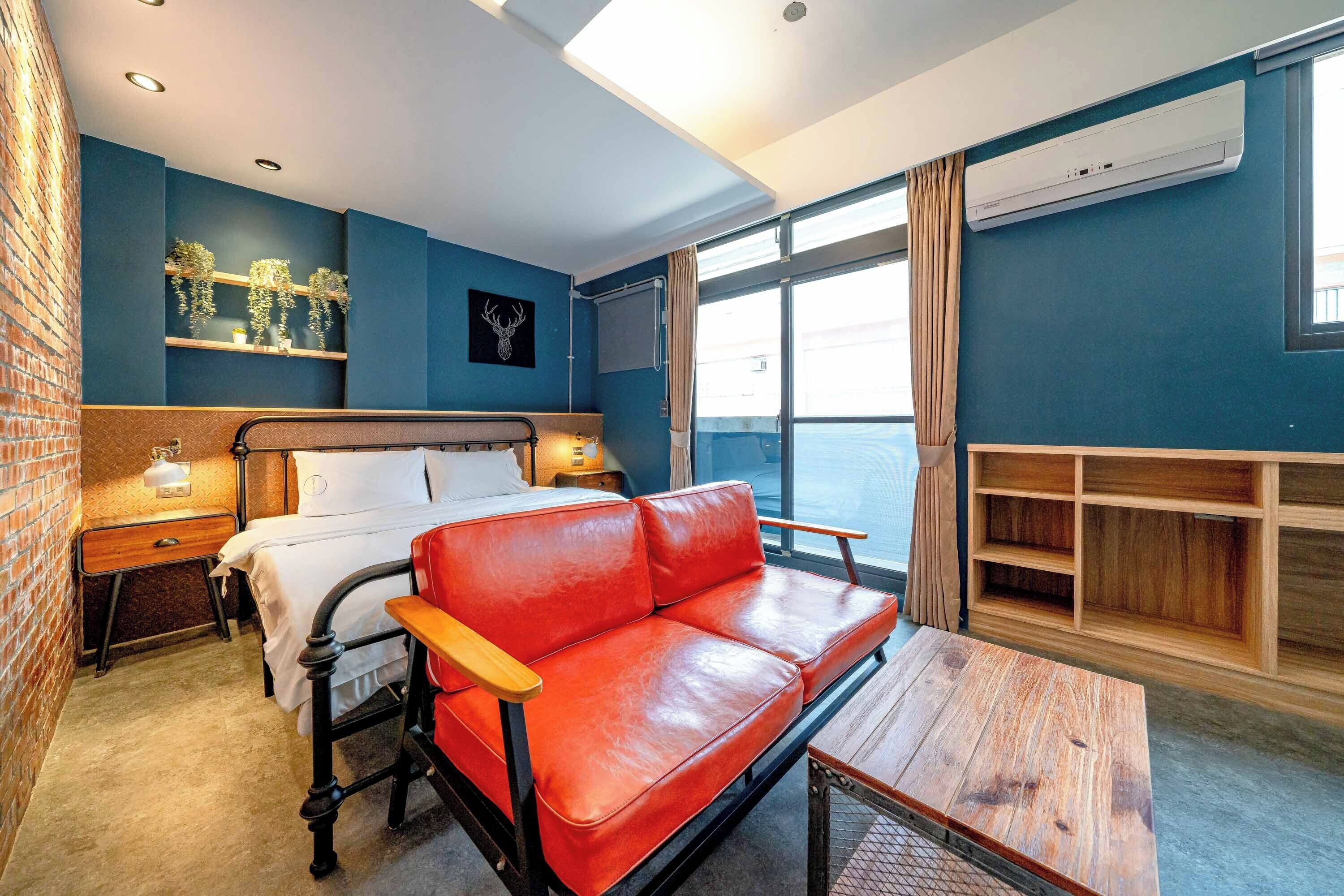 A hotel room with a bed, a red leather sofa, a wooden table, a nightstand, a wall-mounted shelf with plants, and a window with curtains.