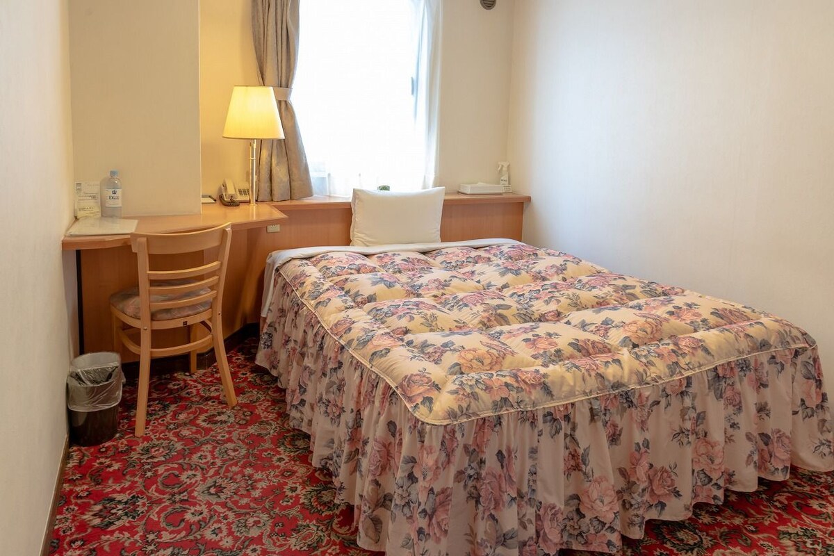 Una habitación de hotel con cama, escritorio, silla, lámpara y ventana con cortinas.