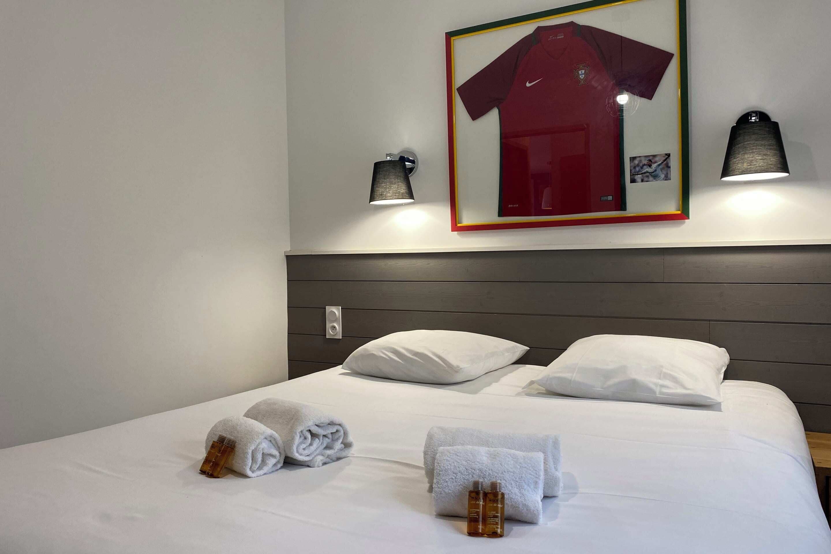 A hotel room with a neatly made bed, two pillows, two rolled towels, and two bottles of toiletries.