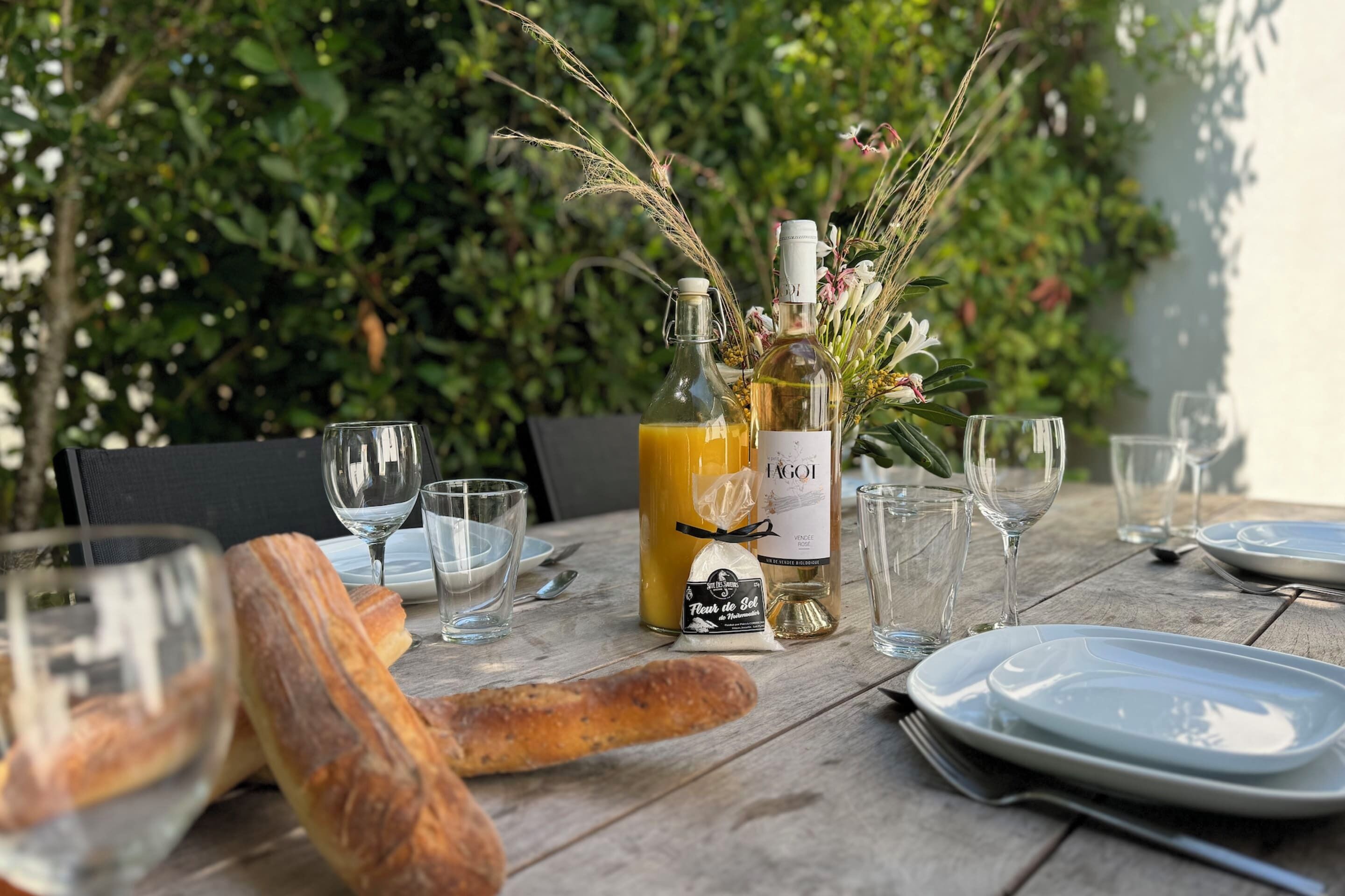 Una mesa de madera dispuesta con platos, vasos, una baguette y botellas de vino, sobre un fondo verde y exuberante.