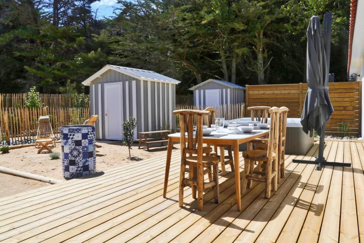 A wooden deck with a table and chairs, a trash can, and two sheds in a backyard.