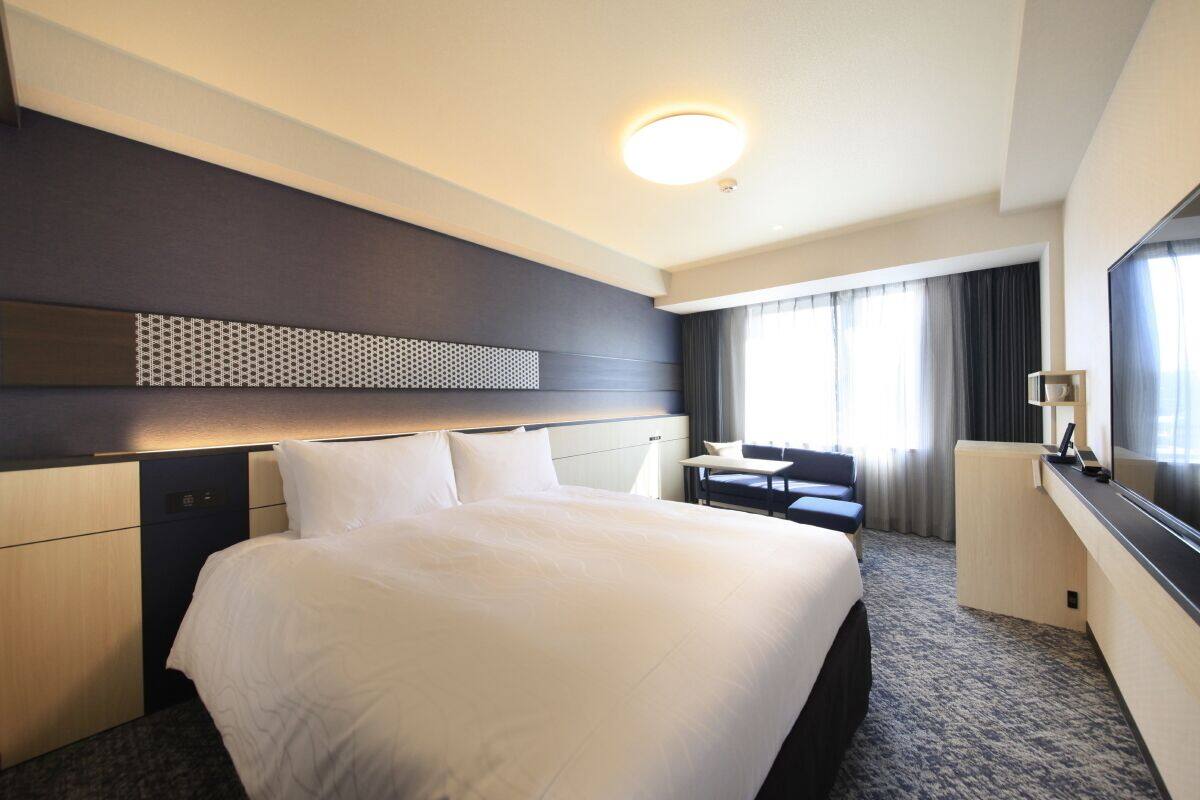 Habitació d'hotel amb un llit gran, una taula de treball, una cadira, una televisió i una finestra amb cortines.