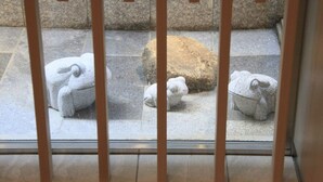 Vista a través de una ventana con marco de madera de tres esculturas de piedra sobre un suelo de baldosas.
