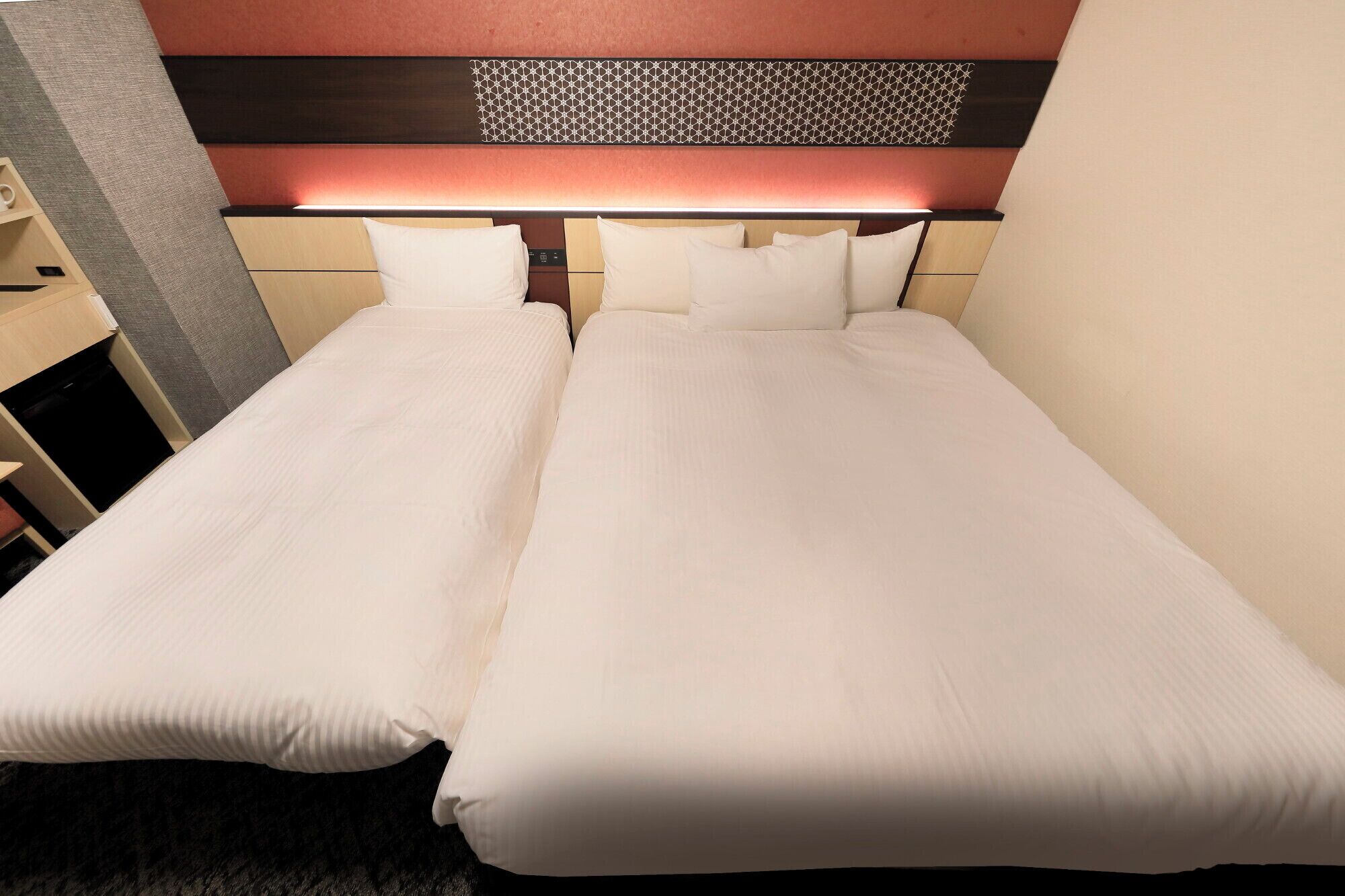 Cama doble con cabecera de diseño geométrico, mesita de noche y lámpara fijada a la pared.