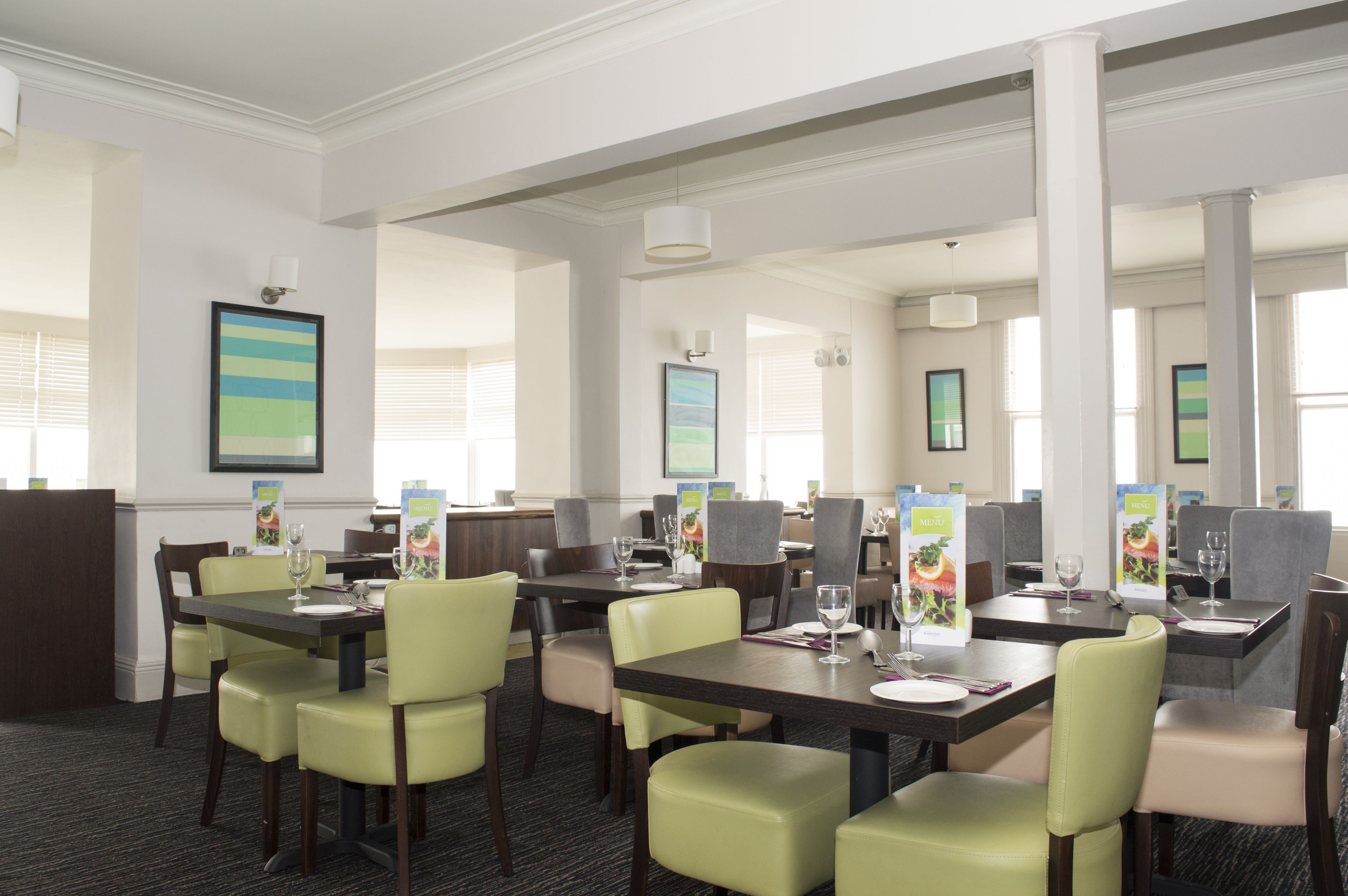A hotel dining area with multiple tables and chairs, framed artwork on the walls, and menu boards.