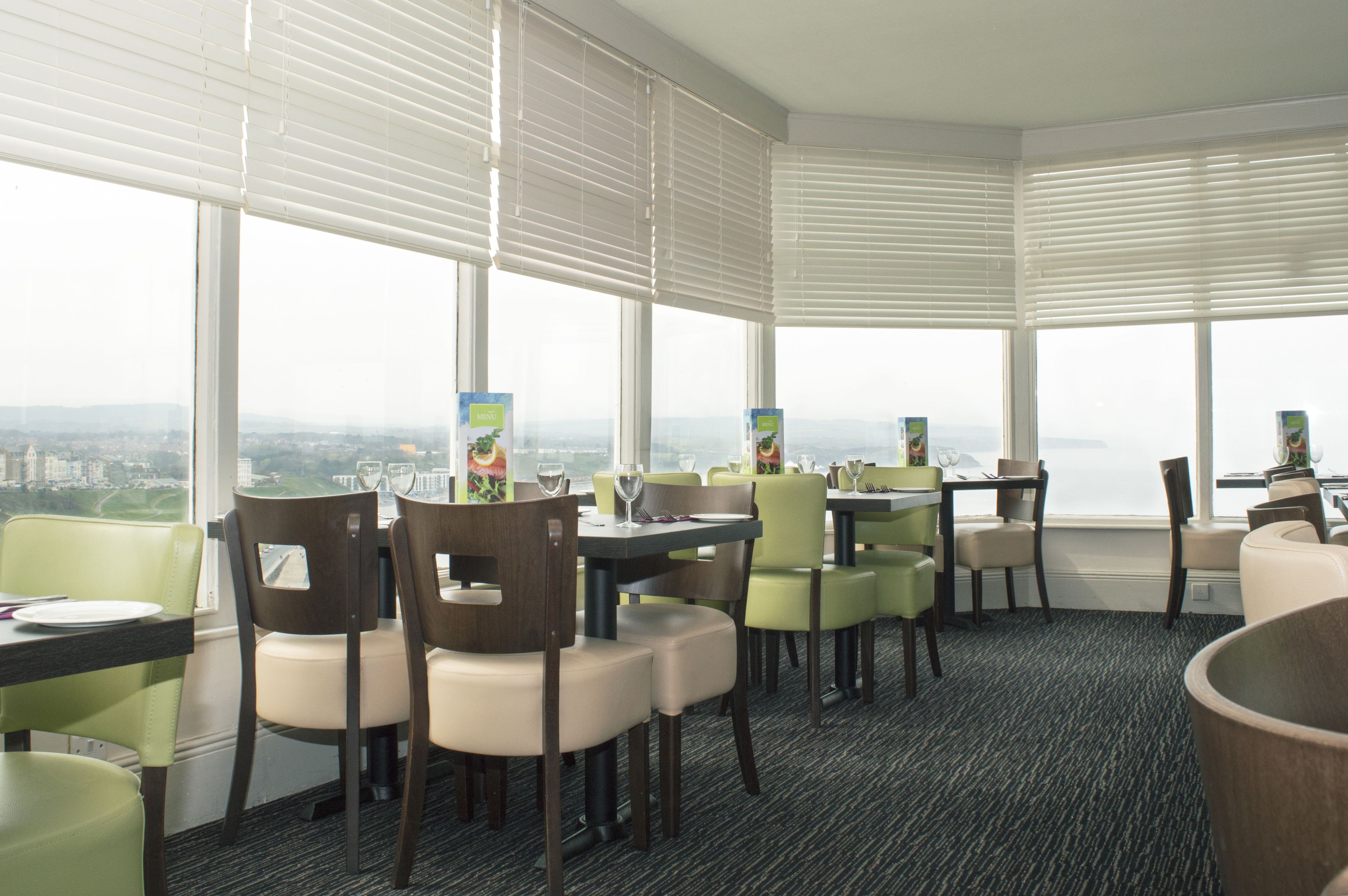 Um espaço de refeições de hotel com várias mesas e cadeiras, amplas janelas com persianas e piso coberto por carpete.